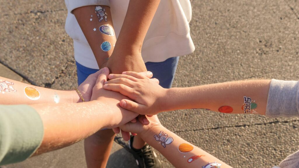 Home Children with space-themed tattoos stack hands symbolizing unity and togetherness.